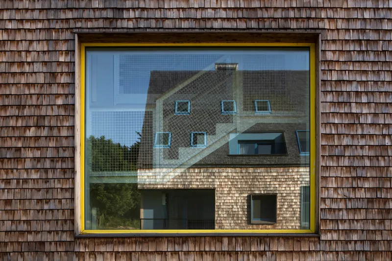 Yellow-framed window in a weathered wood shingle facade reflecting the adjacent shingle-clad building under a clear sky