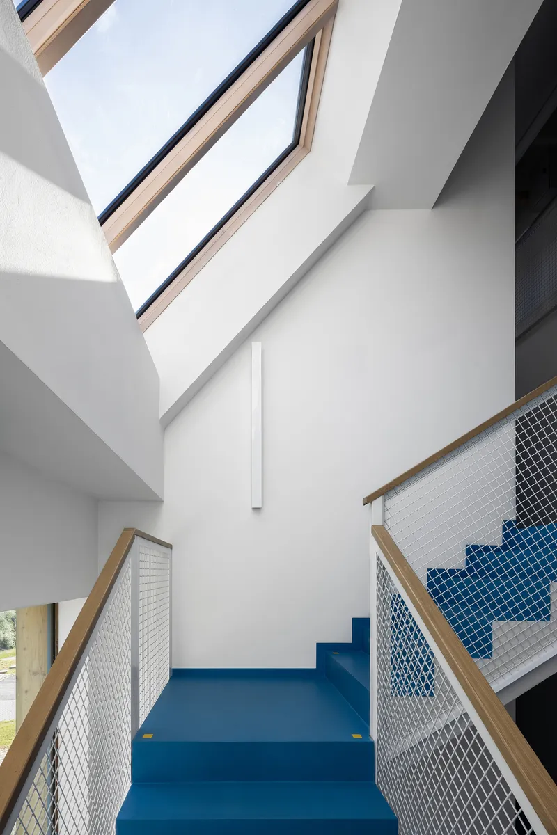 Double-height interior stair with blue rubber flooring, mesh balustrades with timber handrails, and angled skylights above