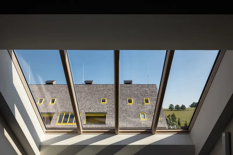 View through angled roof windows framing the grey shingle roof and yellow-trimmed dormers of the neighboring volume