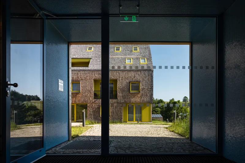 Entrance courtyard framed by dark mesh screens looking toward the shingle-clad facade with yellow window reveals