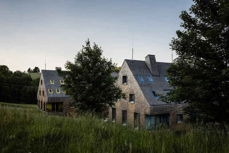 Two gabled volumes with slate roofs and stone cladding set in tall grass at dusk