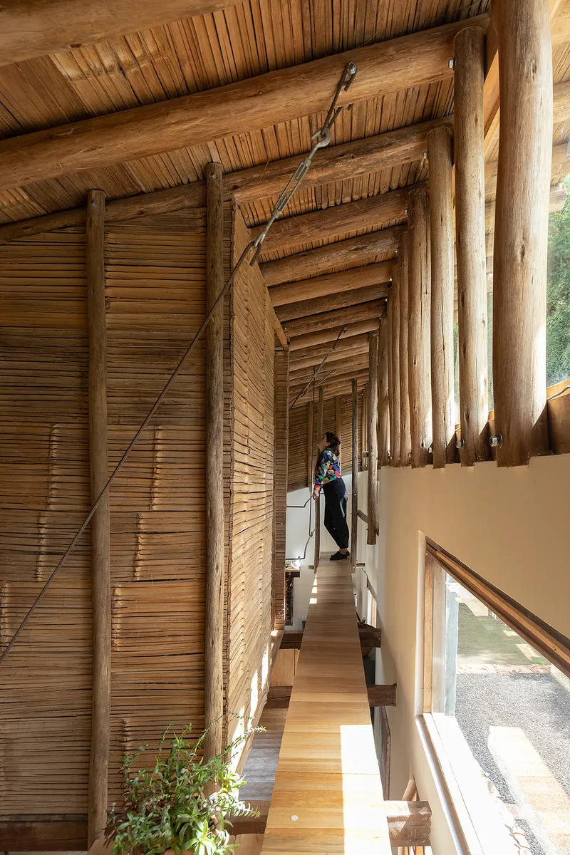 Upper-level timber walkway with bamboo-clad wall, branch bracing, and a person walking in afternoon light