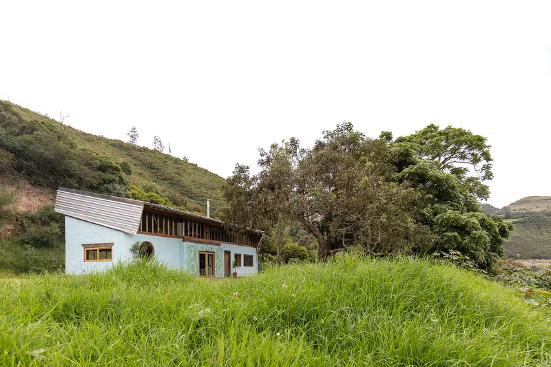 White-painted house with corrugated metal roof nestled into grassy hillside beneath mature trees