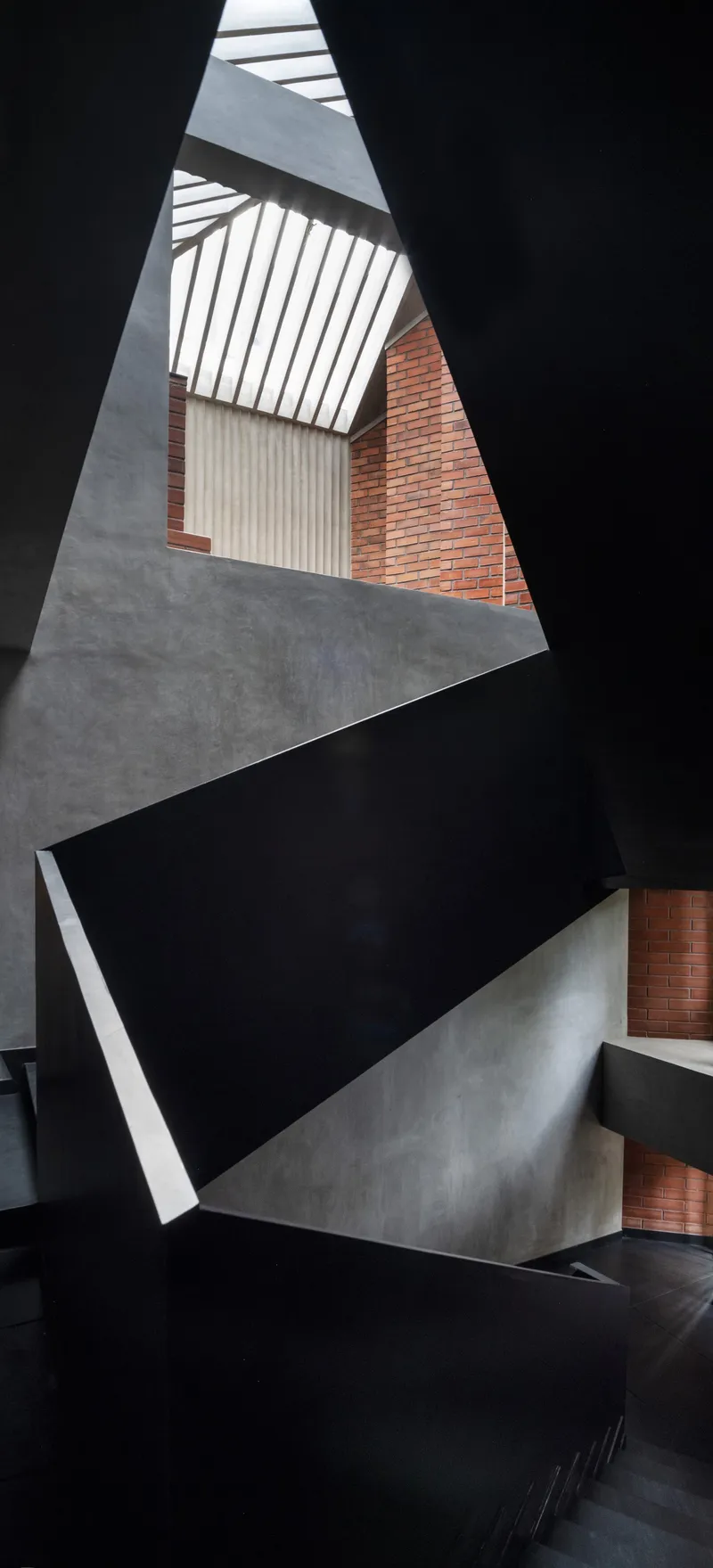 Vertical view up the stairwell showing triangular concrete landings and black steel flights beneath slatted skylight