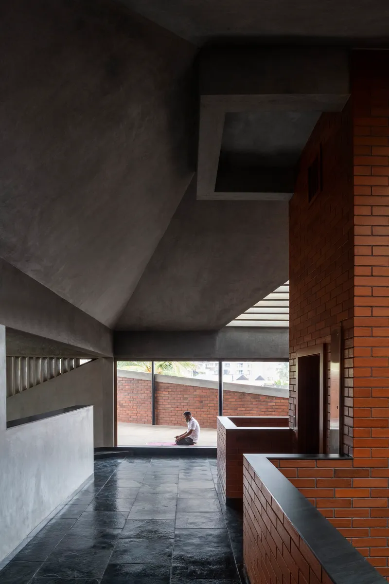 Multi-level interior space with faceted concrete ceiling and red brick volumes with a seated figure by horizontal window