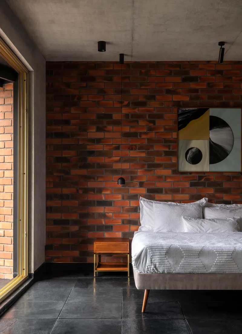 Bedroom with variegated red brick wall, exposed concrete ceiling and dark stone floor beside a glazed opening