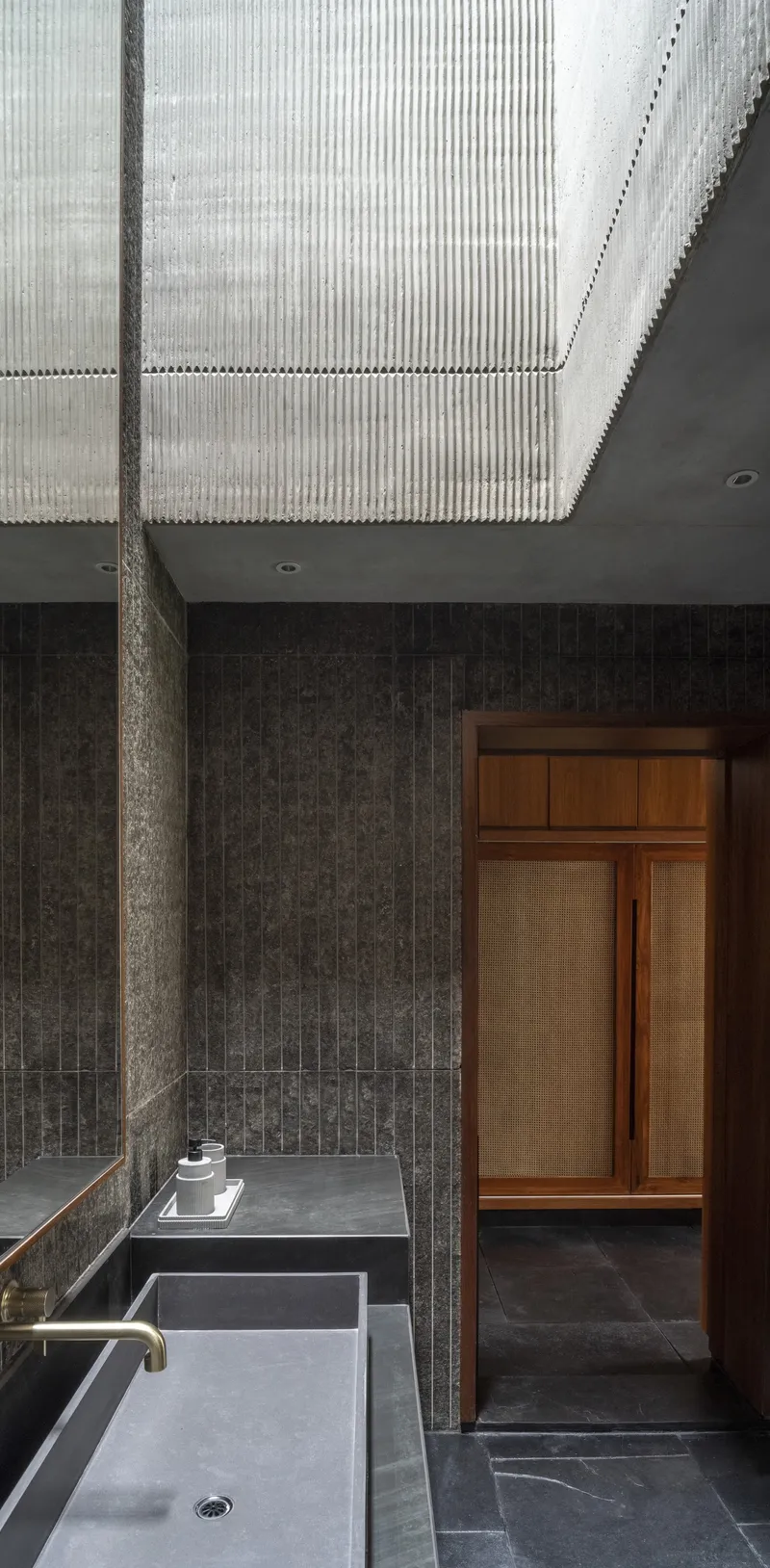 Bathroom with ribbed concrete ceiling above dark brick walls and a sunken grey stone bathtub