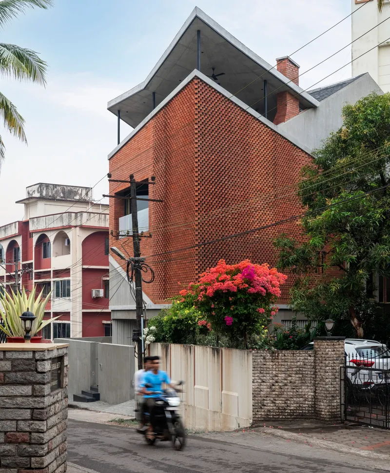 Street view showing the angled brick facade with a motorbike passing and flowering plants at the boundary wall