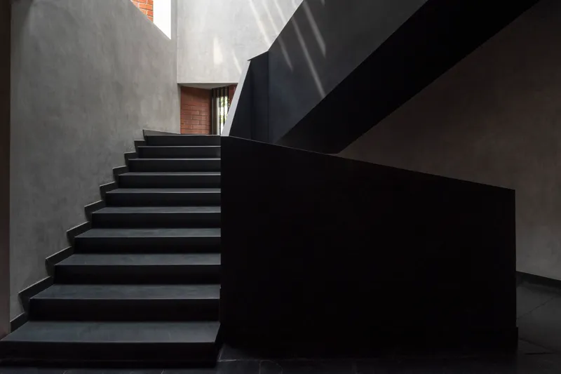 Black cantilevered staircase rising between grey concrete walls with diffused daylight from above