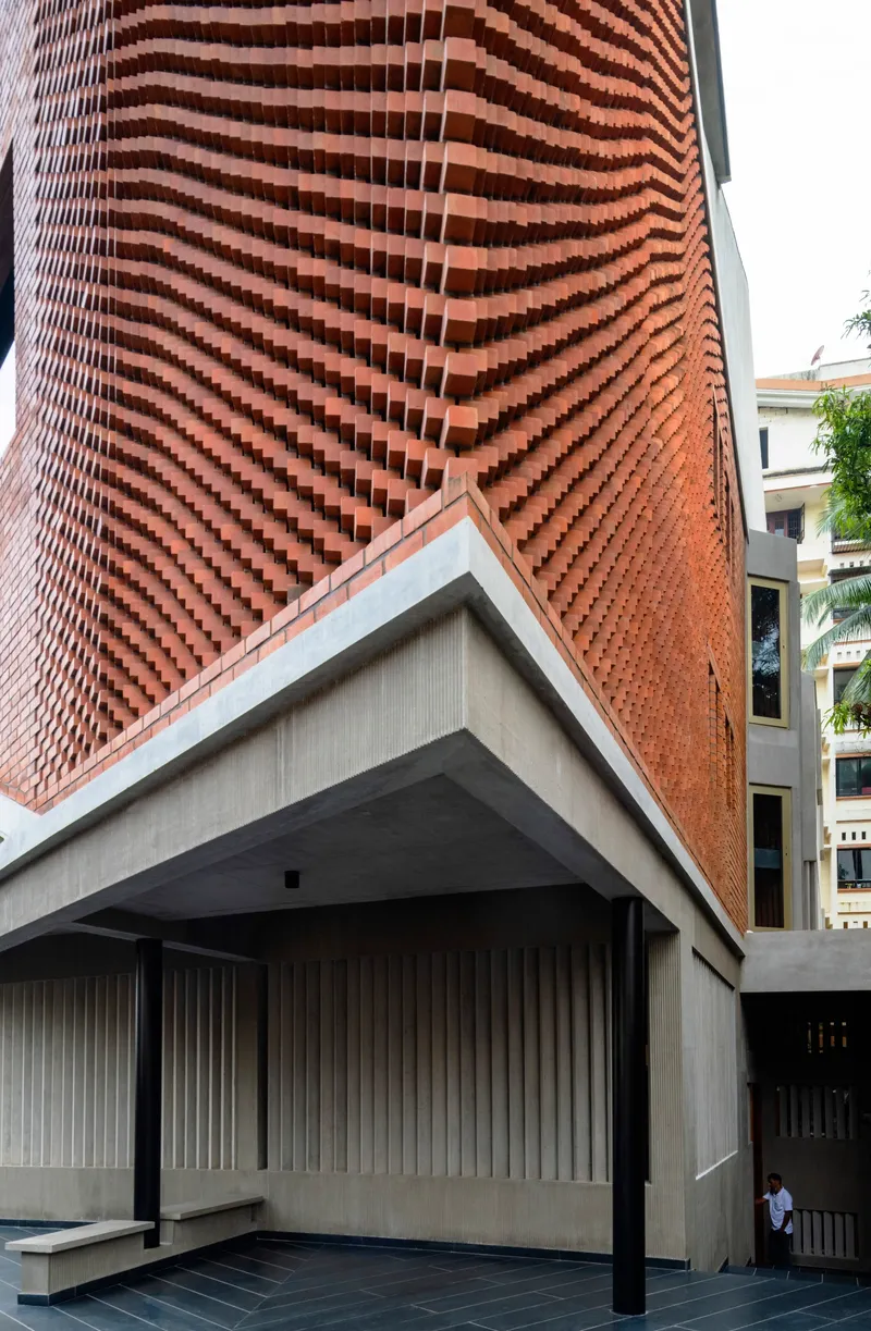 Undulating chequered brick facade meeting angular concrete soffit above a covered entry with vertical timber cladding
