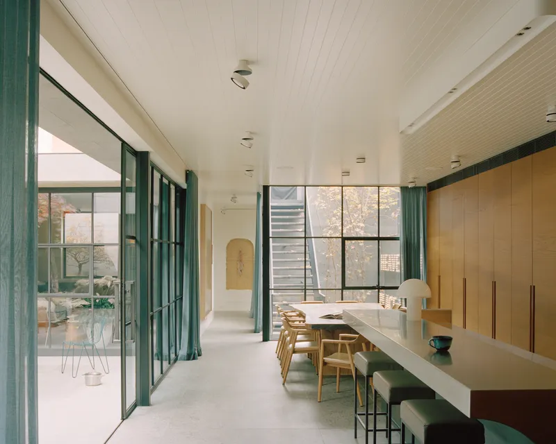 Open-plan dining room with timber cabinetry, steel-framed glazing and green curtains framing a courtyard