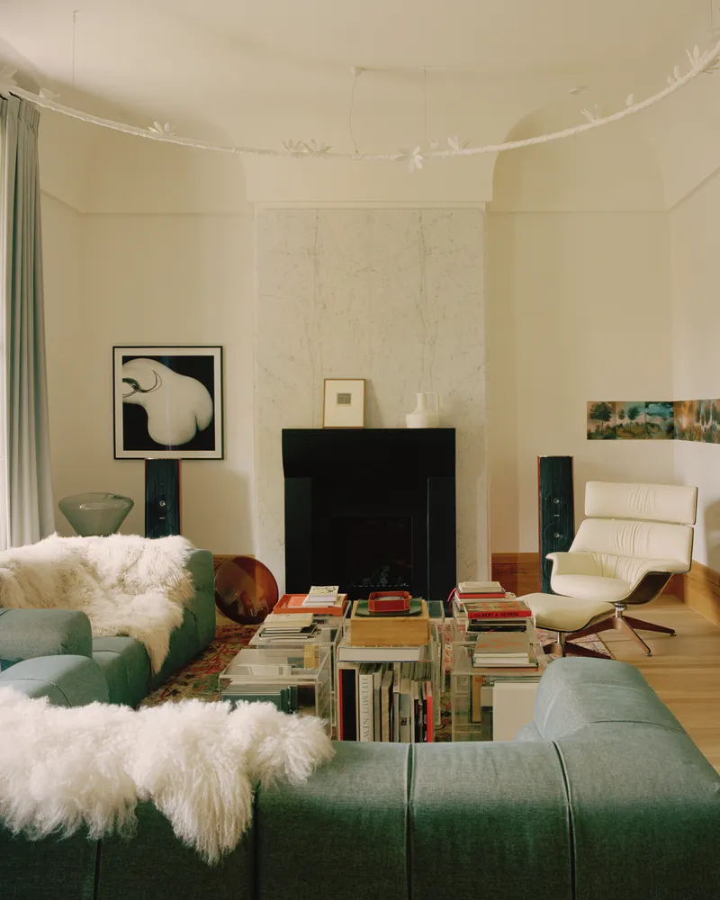 Living room with black fireplace surround, acrylic book tables and green sectional sofas with sheepskin throws