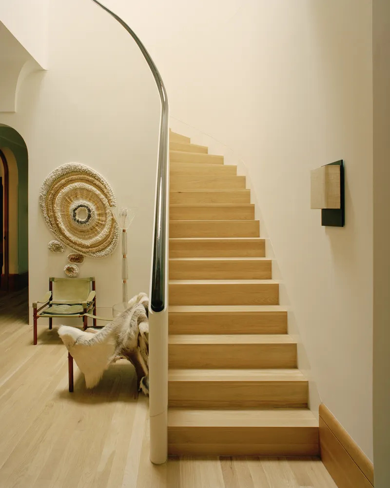 Oak staircase with curved steel handrail ascending beside white walls and a chair with woven wall hanging