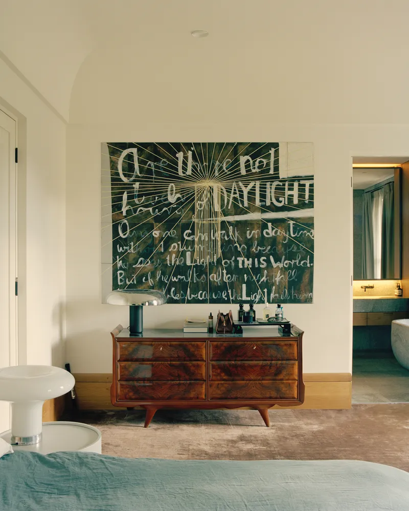 Bedroom with walnut dresser beneath text-covered artwork and doorway opening to an ensuite bathroom