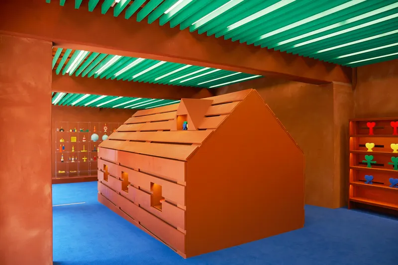 House-shaped timber storage structure with slot openings beneath green slatted ceiling beams and blue rubber flooring