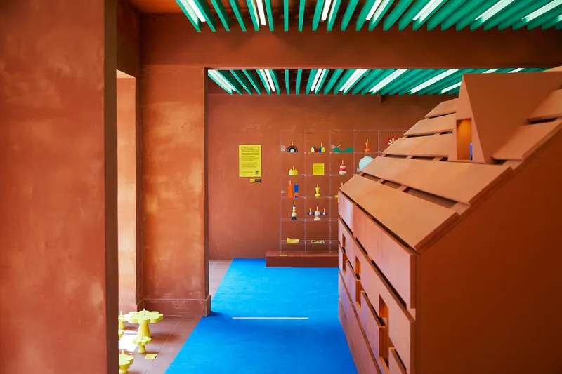 Interior passageway with ochre plastered walls, angled timber stair, and green ceiling slats filtering daylight