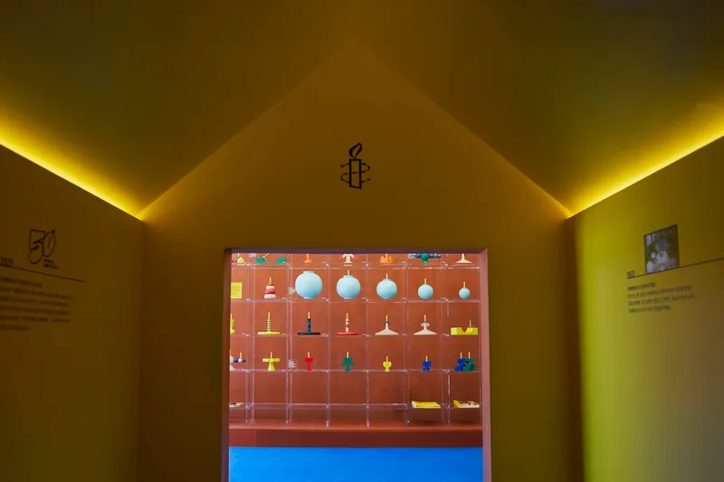 Doorway framing a display vitrine with suspended objects, yellow cove lighting illuminating the gabled ceiling