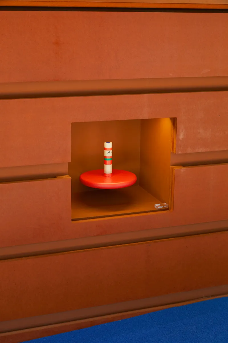 Square niche in terracotta-toned wall with a red round shelf and white candle inside