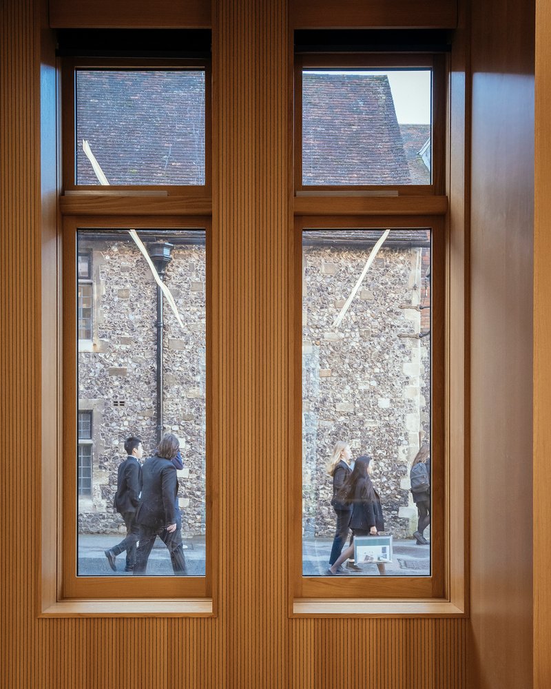 Interior view through timber-framed windows showing pedestrians passing weathered stone walls outside