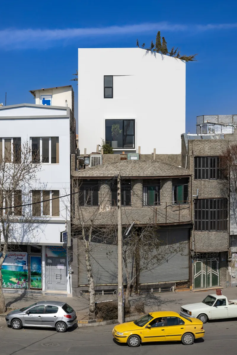 Street view of a white cubic volume rising above neighboring brick structures with a yellow taxi passing