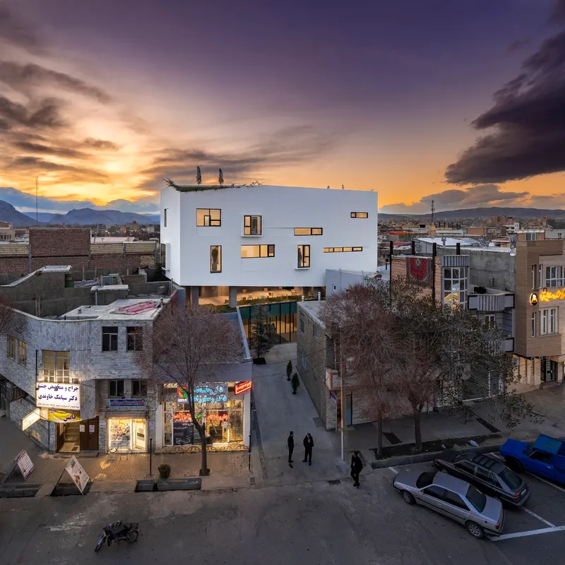 White volume with illuminated windows at dusk above pedestrians and storefronts in the street below
