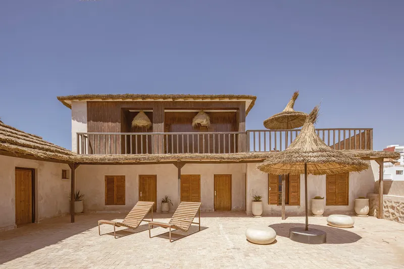 Two-story facade with timber shutters and upper balcony overlooking terrace with lounge chairs and thatch umbrellas