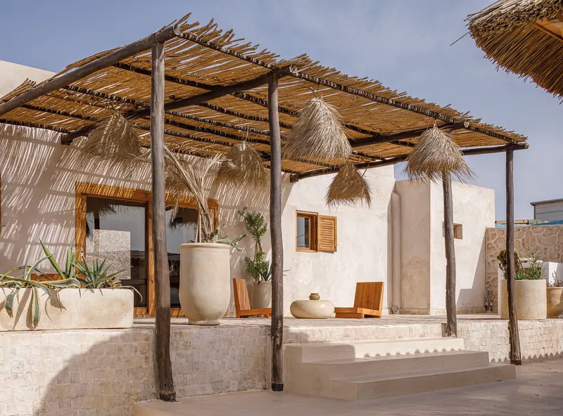 Outdoor pergola structure with bundled reed canopy, timber columns, hanging thatch shades, and planted ceramic vessels on stone platform