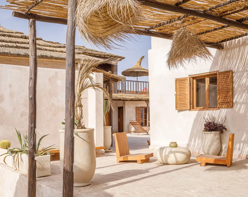 Courtyard view through timber pergola posts showing whitewashed walls, timber shutters, planters, and upper balcony with thatched parasol
