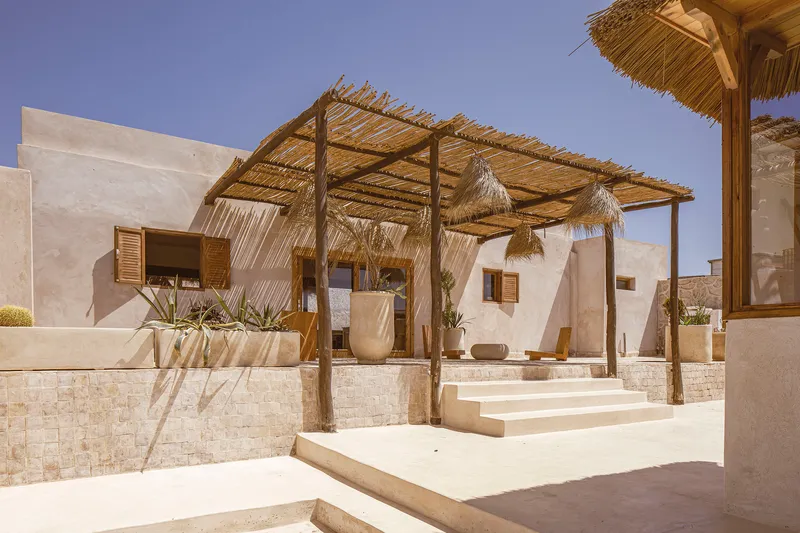 Exterior terrace with timber pergola and thatched roof over stone planters and steps in bright sunlight