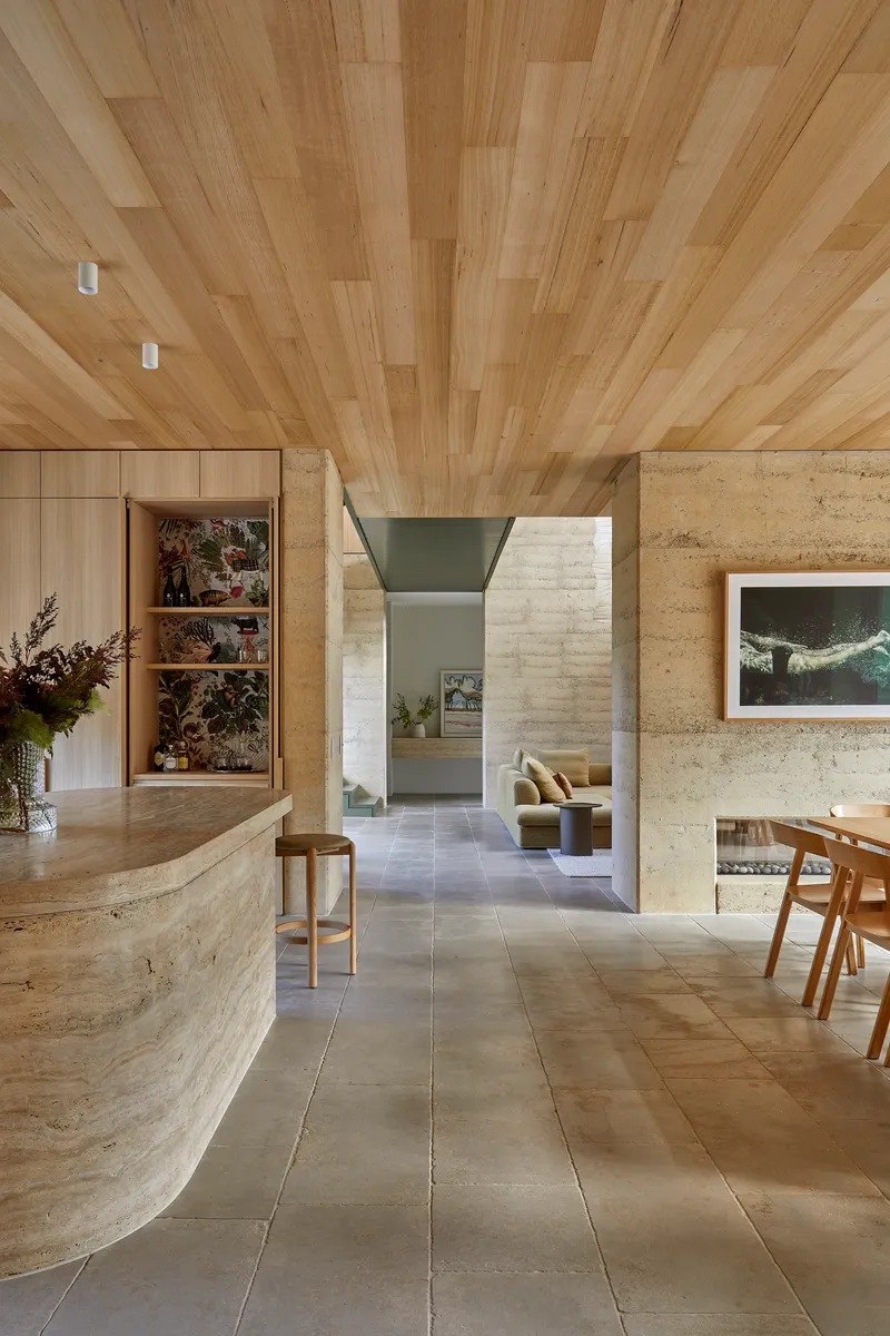 Open plan living space with timber plank ceiling and curved rammed earth kitchen island