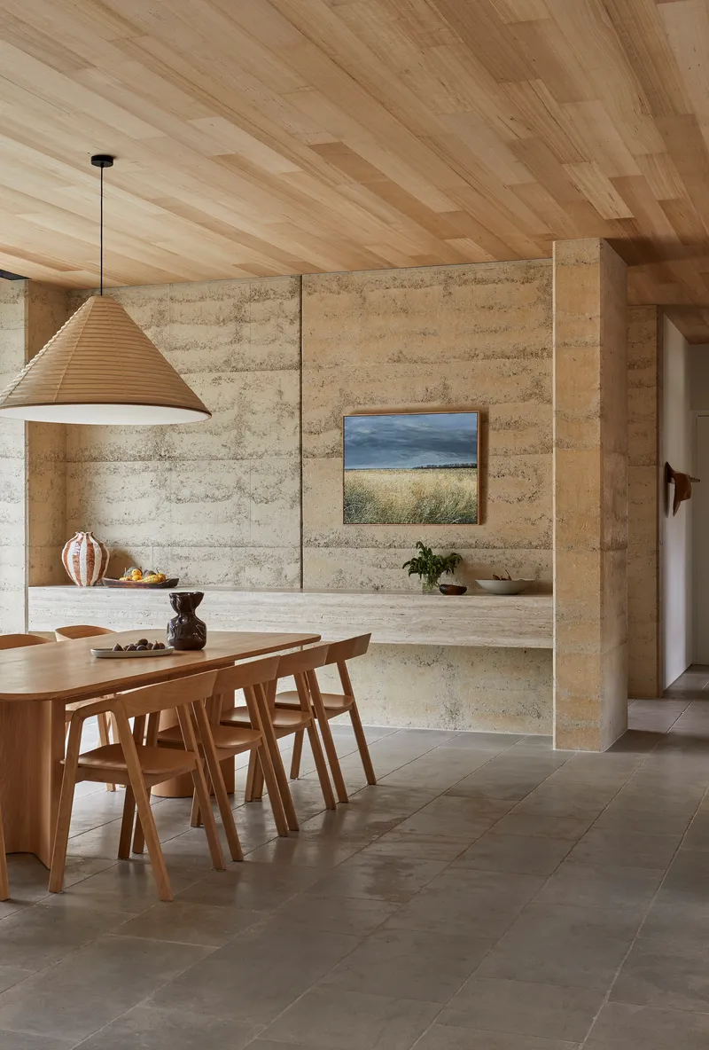 Dining area with rammed earth walls, timber plank ceiling and large pendant light above an oval table
