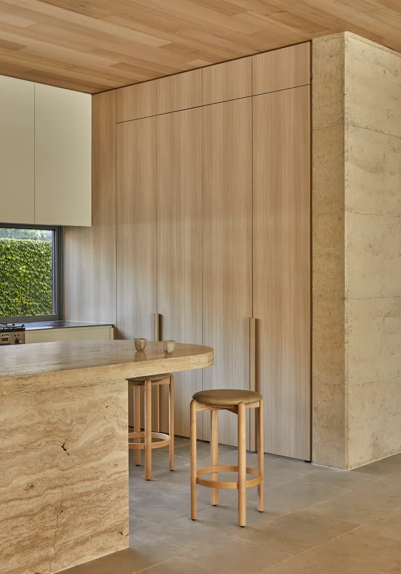 Kitchen with vertical grain timber cabinetry beside a rammed earth breakfast bar and two stools