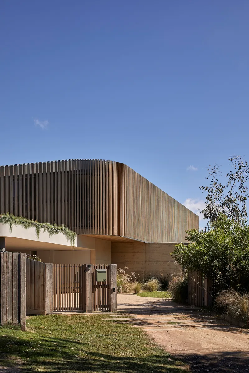 Curved timber-clad upper volume above rammed earth base with slatted gate and lawn