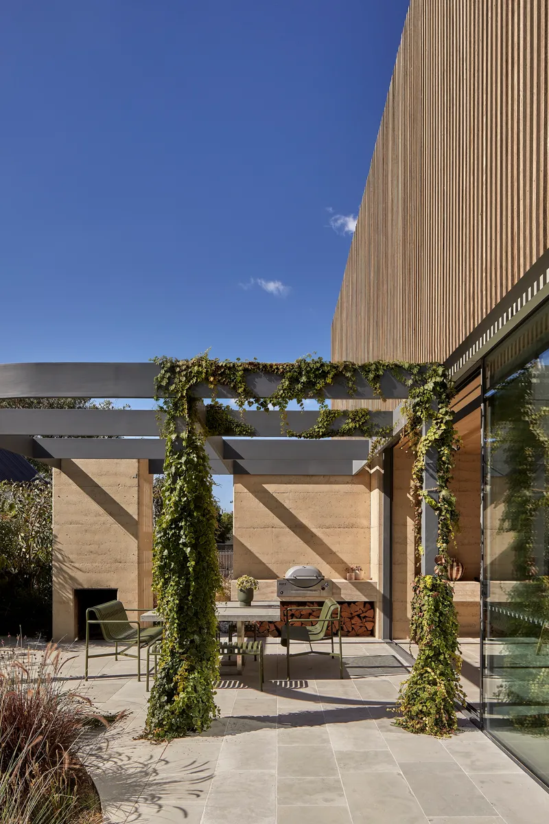 Outdoor terrace with vine-wrapped steel pergola beneath vertical timber-clad upper volume