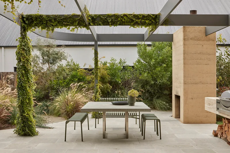 Outdoor dining terrace beneath a steel pergola draped with climbing vines and planted garden beds