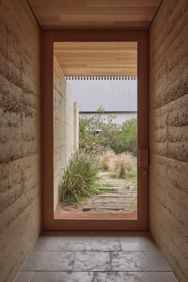 Glazed timber door framed by rammed earth walls opening to a gravel path and grasses