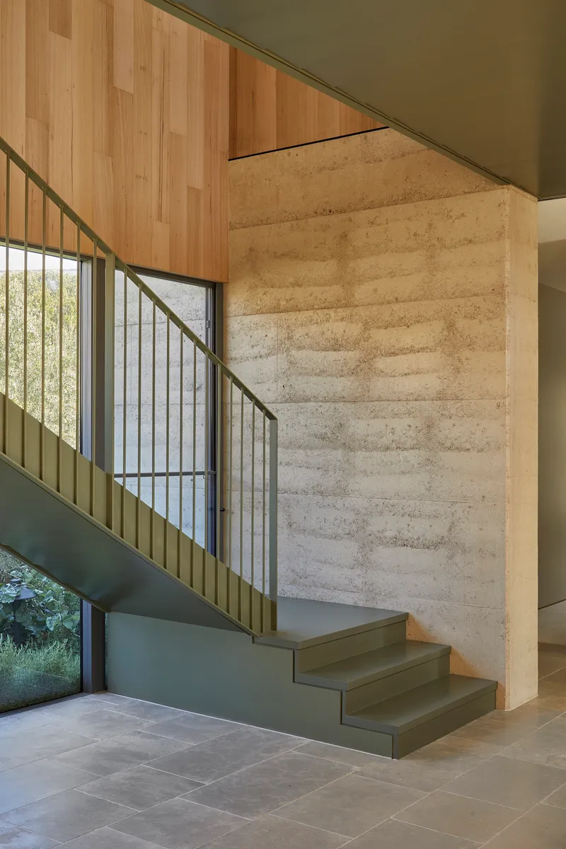 Staircase with metal balustrade rising beside a rammed earth wall and timber ceiling above