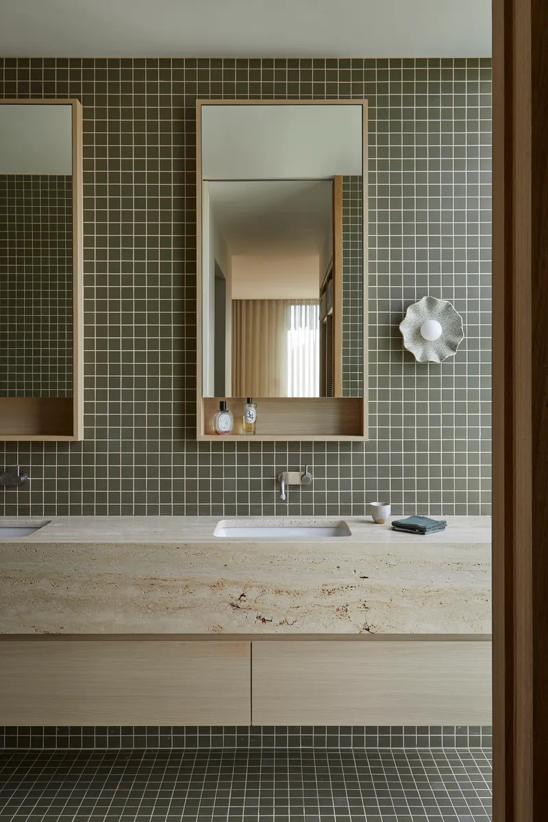 Bathroom vanity with olive green mosaic tile and travertine countertop below framed mirrors