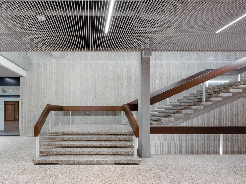 Terrazzo staircase with angled timber handrails set against ribbed concrete walls beneath a slatted wood ceiling