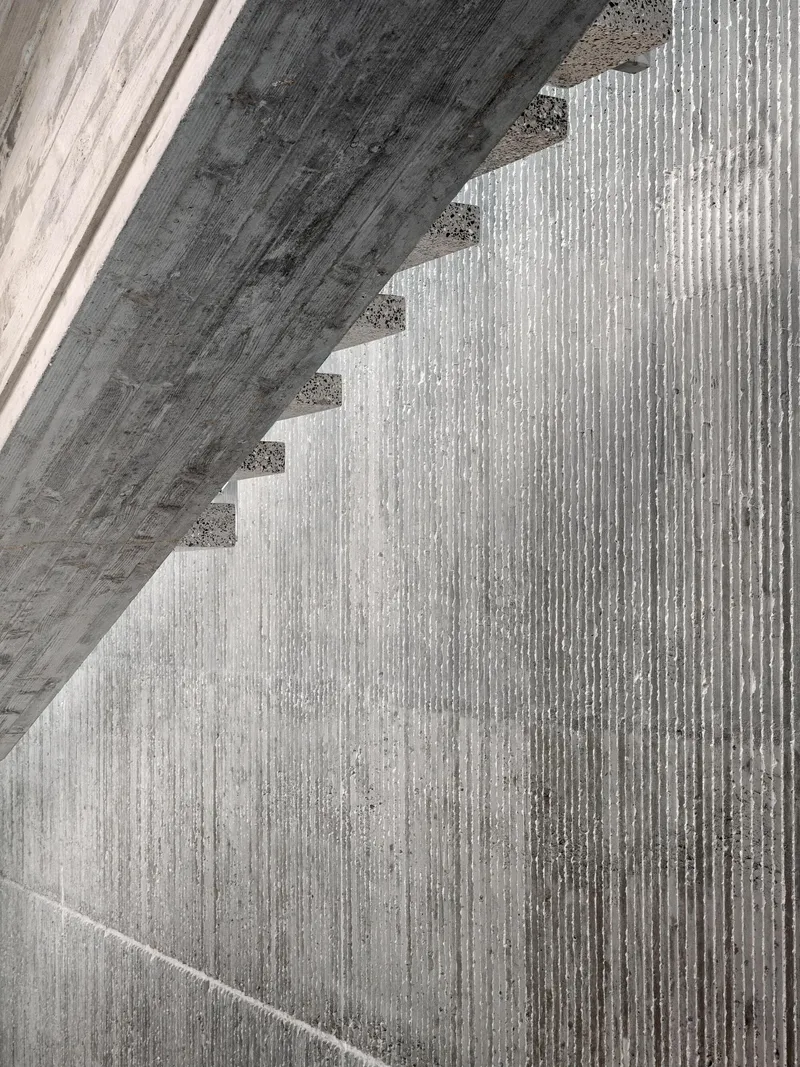 Close-up of board-formed concrete stair soffit casting diagonal shadows across ribbed wall