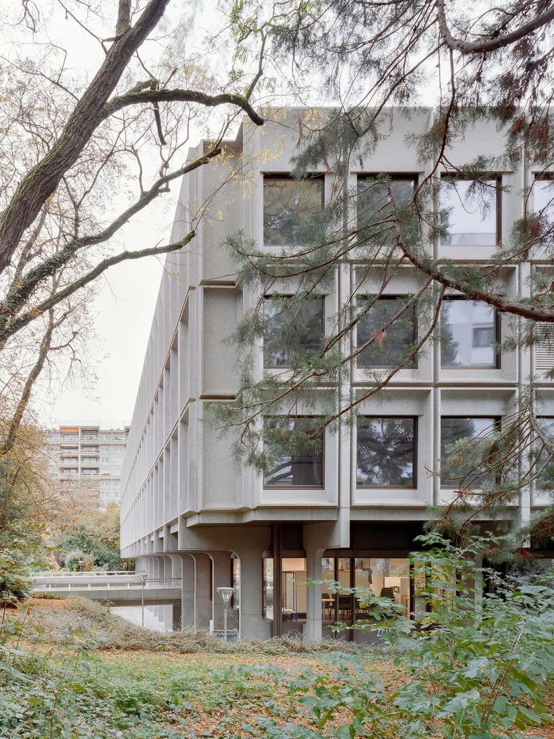 Elevated concrete building on piloti viewed through bare winter trees and undergrowth