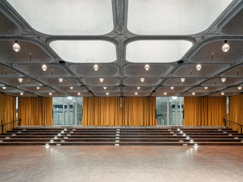 Auditorium interior with exposed concrete waffle ceiling, pendant lights, and mustard curtains