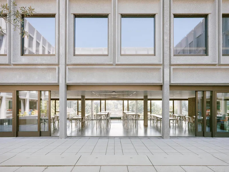 View across the courtyard terrace toward the dining hall with floor-to-ceiling windows and suspended ceiling panels