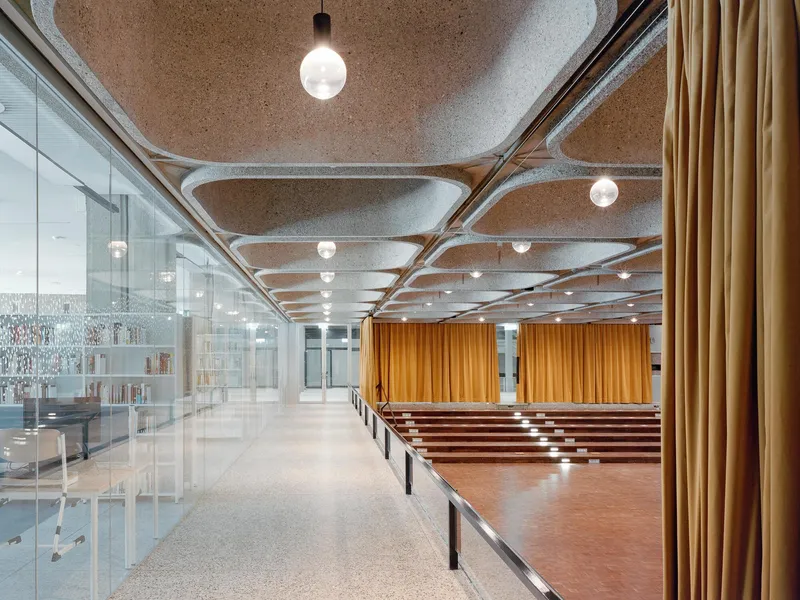 Interior foyer with cork ceiling panels, white terrazzo floors, glass partition and tiered wooden seating under pendant lights