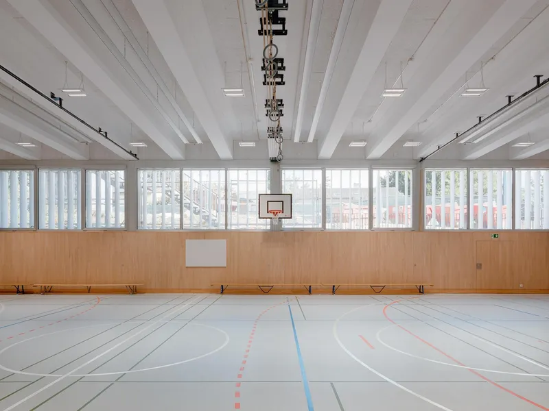 Gymnasium with painted floor lines, basketball hoop, exposed white ceiling beams and translucent perimeter screens