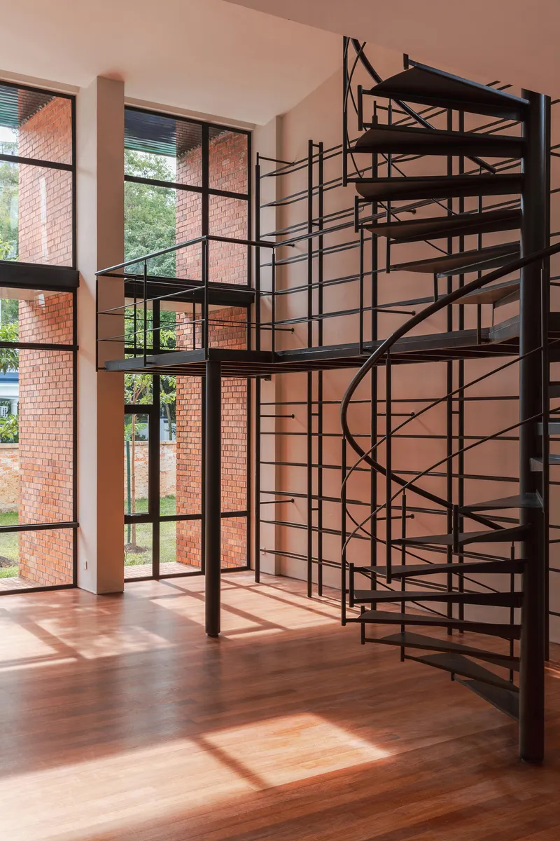 Black steel spiral staircase beside floor-to-ceiling windows overlooking brick courtyard with dappled sunlight on timber floor