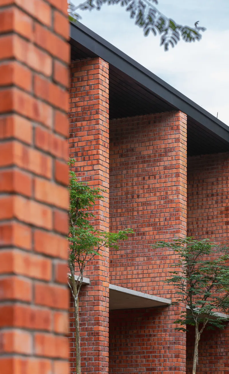 Red brick facade with stacked square columns beneath a dark roof overhang and tree branches
