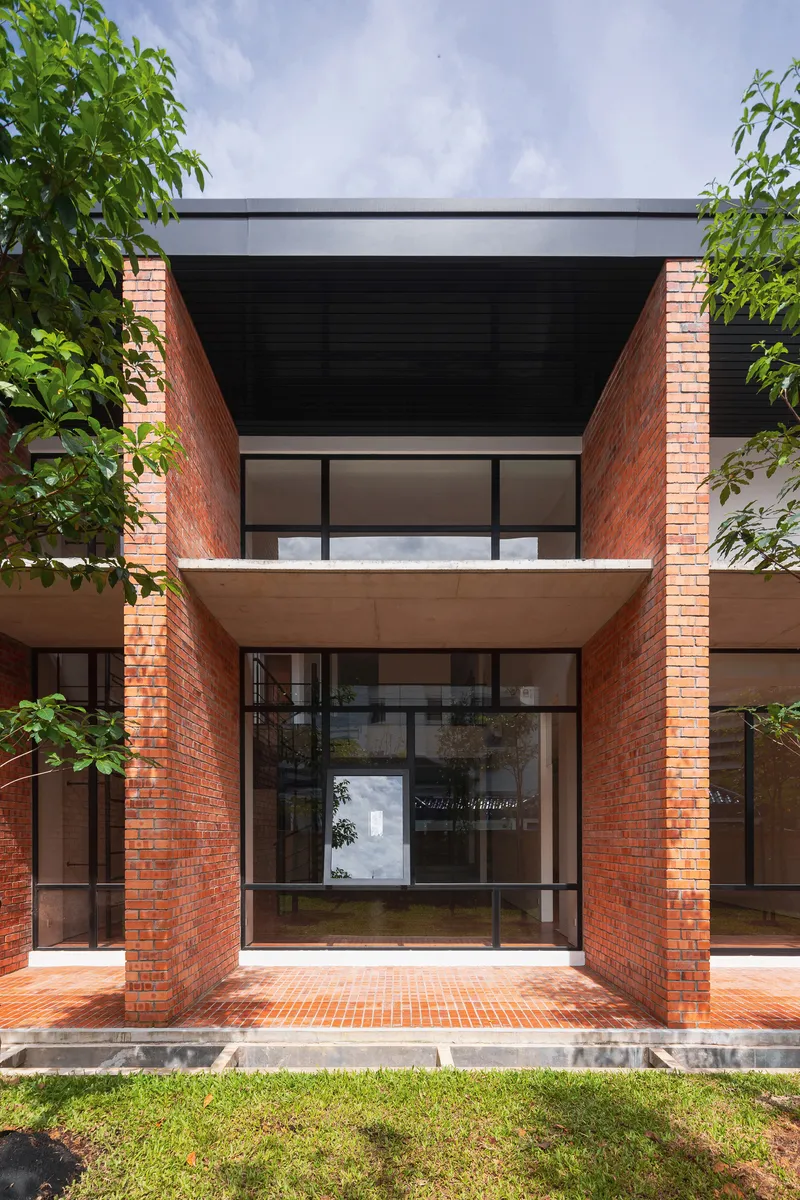 Front facade with red brick columns framing a glazed entrance and recessed upper balcony under lush green trees