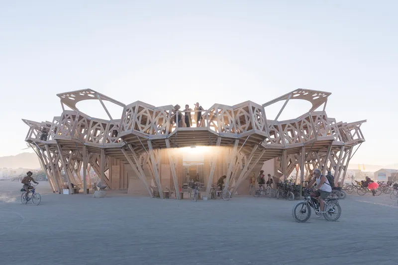 Latticed timber pavilion with patterned apertures and upper deck as cyclists pass at dusk