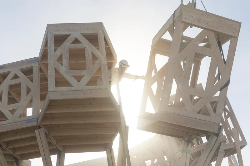 Person climbing between two geometric timber modules with diamond lattice patterns backlit by sun